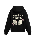 #siyah renk hoodie arka görünüm; sırt baskısında “broken hearts” yazısı, pembe kalp gözlü iki krem kafatası illüstrasyonu ve pembe parıltı ikonları.