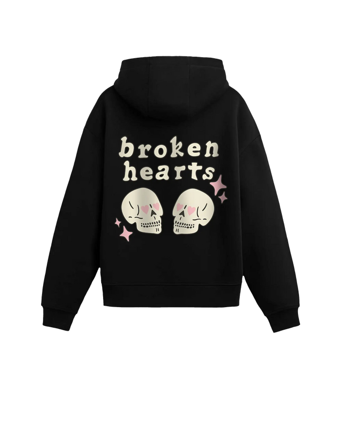 #siyah renk hoodie arka görünüm; sırt baskısında “broken hearts” yazısı, pembe kalp gözlü iki krem kafatası illüstrasyonu ve pembe parıltı ikonları.